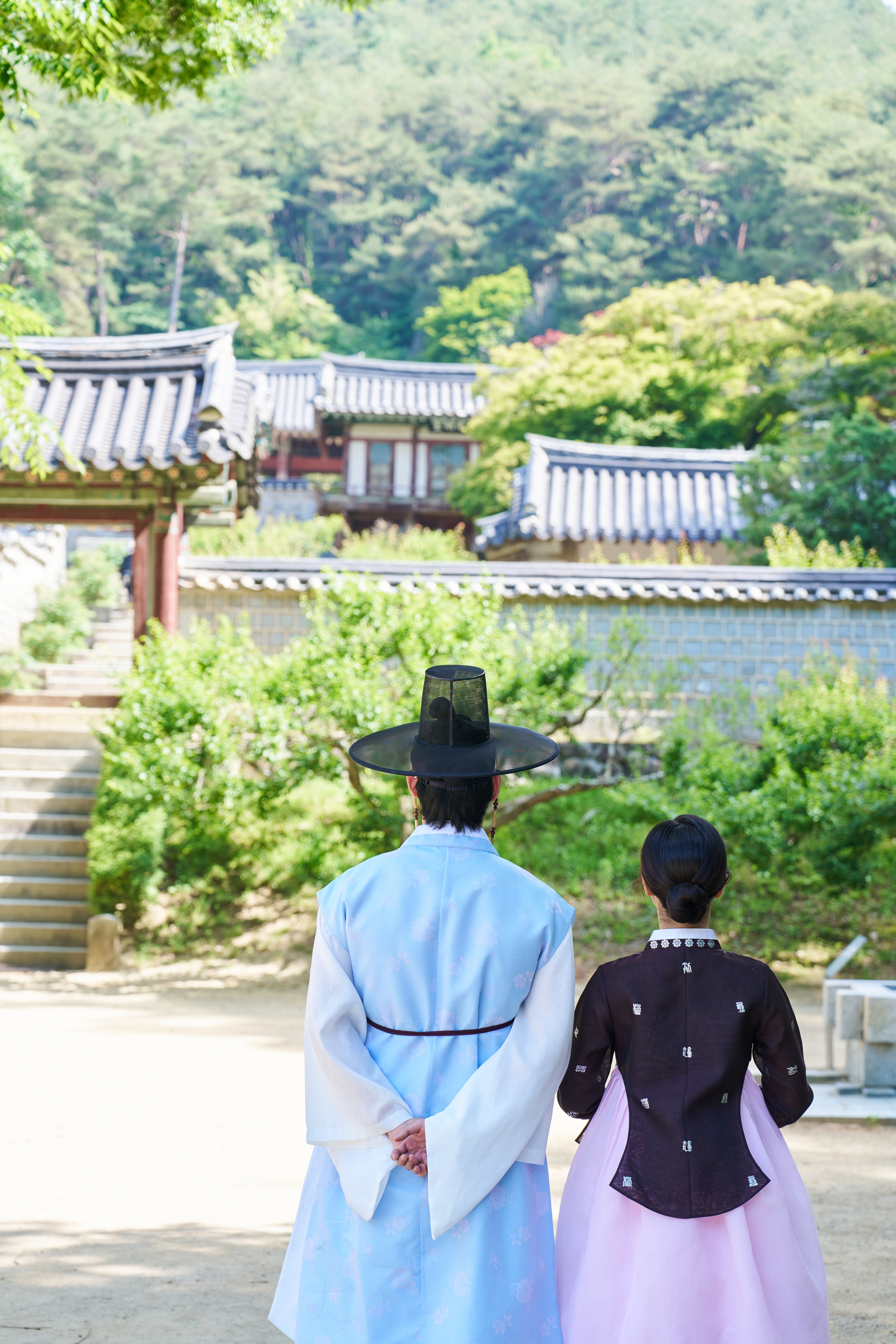 하늘색 두루마기와 검은 갓, 남색 저고리와 연보라 치마 차림의 두 사람이 한옥 지붕과 초록 언덕을 바라보는 뒷모습