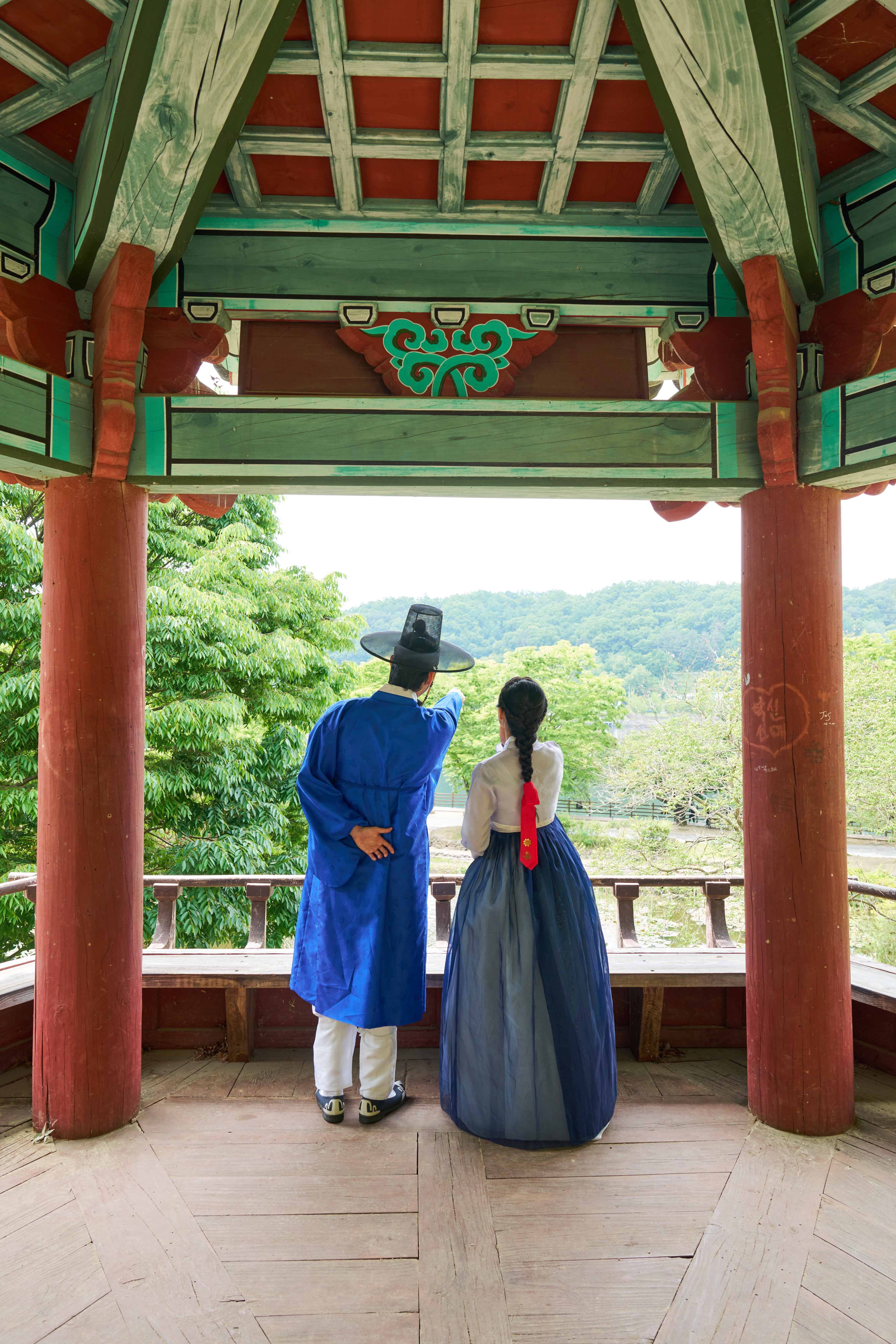 붉은 기둥과 단청 처마 아래 정자에서 초록 풍경을 바라보는 코발트·남색 한복 차림의 두 사람 뒷모습