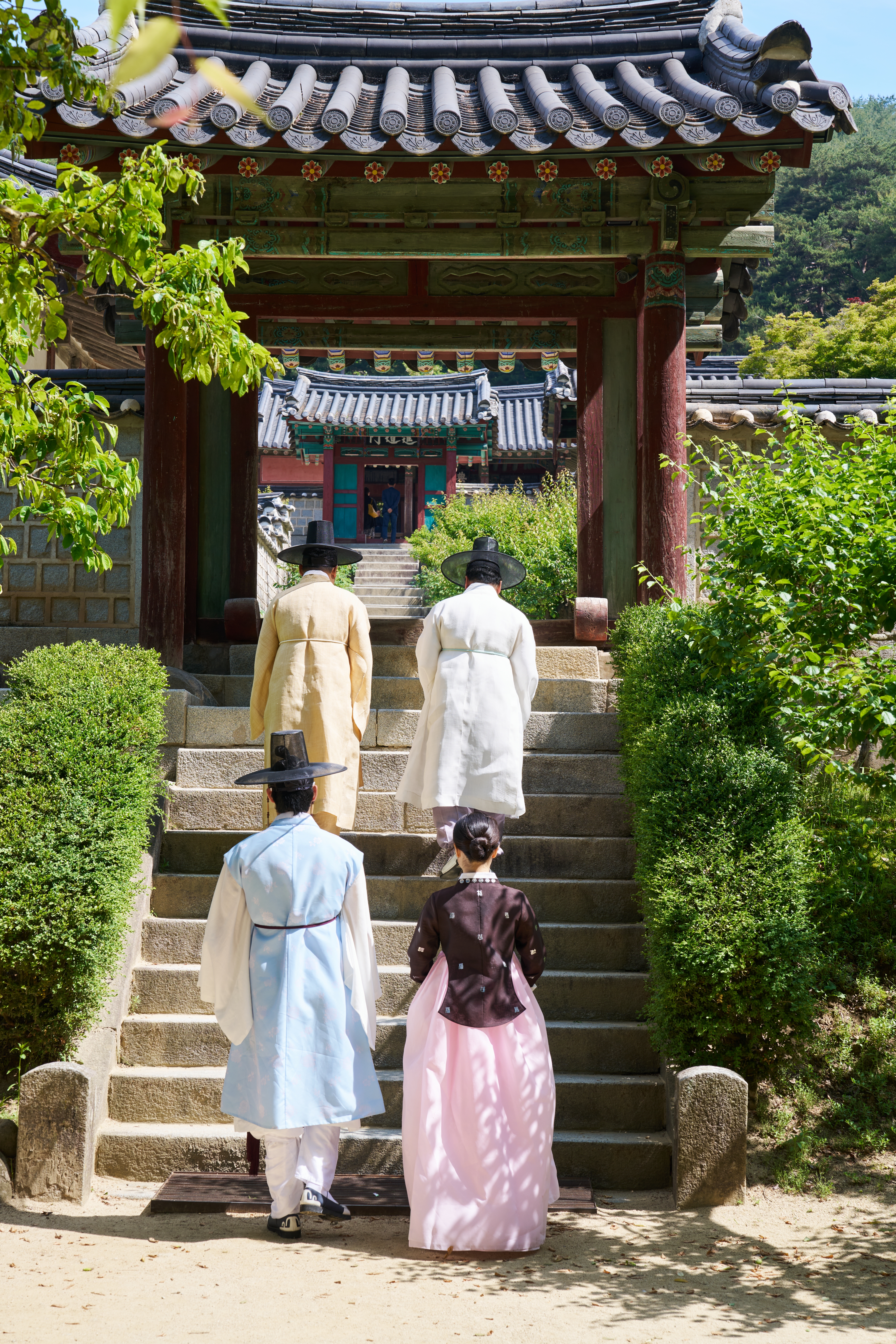 초록 숲 사이 돌계단을 올라 문으로 향하는 한복 차림의 사람들
